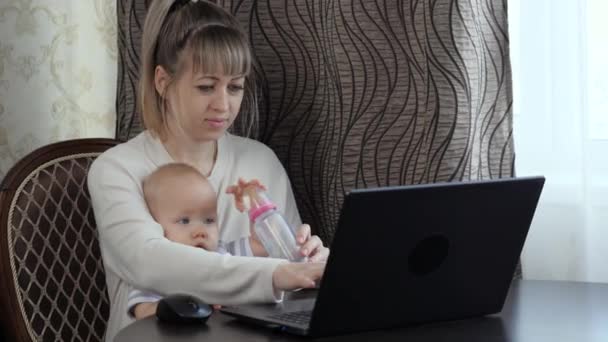 A mãe trabalha no computador com a criança. O miúdo senta-se com a mãe, bebe, come de uma mamadeira. Mulher freelance com criança. Mulher de negócios trabalhando em casa no laptop com criança à mesa — Vídeo de Stock