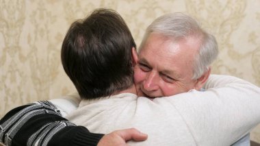 Mutlu gri saçlı baba, evde kucaklaşan yetişkin oğluyla tanıştığı için mutlu. Ailecek keyifli bir toplantı. Oğlu babasına sarıldı. Kollarında yetişkin bir oğlu olan yaşlı bir baba. Nesiller boyu. İki erkek sarılıyor..
