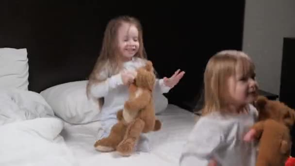Las pequeñas hijas juegan en el sofá por la mañana con su oso de juguete favorito, se divierten, tontean. Niños, niñas, hermanas, salten en pijama en la cama en la habitación y ríanse alegremente. Feliz infancia en casa — Vídeos de Stock