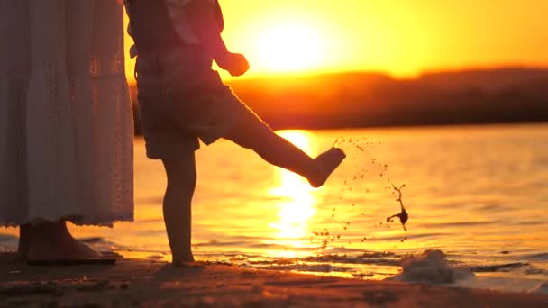 Mother with her daughter on beach, child is playing with her little foot splashing in water in sunshine. Happy family, mom walks with child barefoot on water, holding hands in rays of beautiful sunset — Stock Video