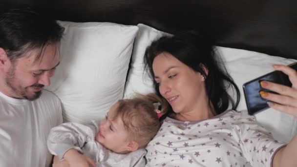 Šťastná mladá rodina s malou holčičkou, jsou fotografovány na smartphone v posteli. Matka, otec a dcera-bloggeři v místnosti komunikovat pomocí moderní gadget on-line. Moderní mladá rodina — Stock video