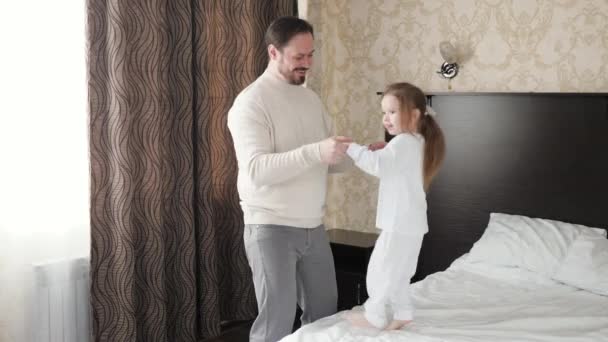 幸せな家族の子供、娘とお父さん、手をつないで、ベッドの上でジャンプします。お父さんは元気に笑い、かわいい娘さんと、寝室で子供と積極的なゲームをしています。朝のダンス。お父さん休み — ストック動画