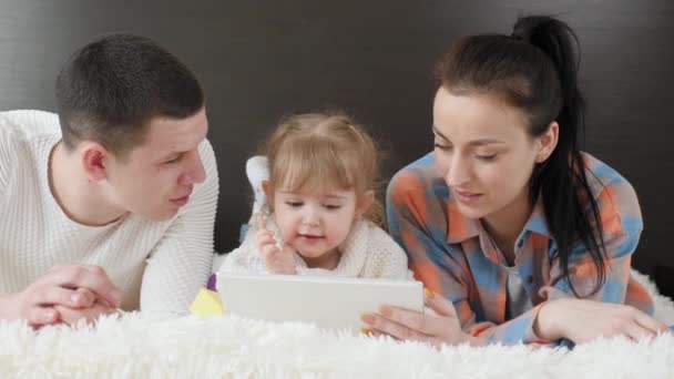 Famille heureuse, parents et petit enfant, fille utilisent la tablette ensemble, couché sur le lit. Mère, enfant et père se reposent le week-end ensemble à la maison. gadget moderne, les parents et les enfants regardent bande dessinée — Video