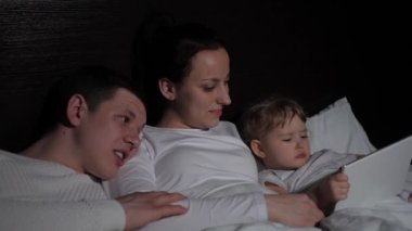 Küçük çocuk, kız dijital tablet kullanarak çizgi film izliyor, ebeveyn kontrolüyle eğleniyor, yatakta oturuyor. Mutlu aile akşamları evde tabletle birlikte dinleniyor. Çevrim içi öğrenim