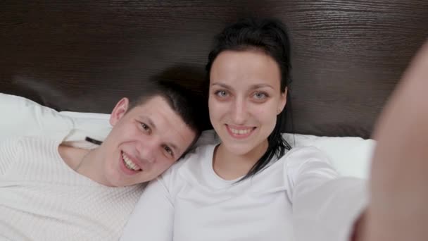 Un jeune couple tenant un smartphone à la main est photographié ensemble, tourne un blog vidéo sur le lit. Heureux mari et femme de famille prendre des photos en ligne à l'aide d'un téléphone portable moderne à la maison sur le canapé dans la chambre. — Video