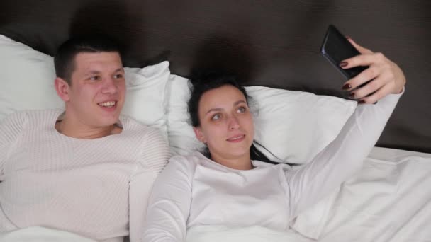 Et ungt par, der holder en smartphone i hånden, fotograferes sammen, skyder videoblog på sengen. Glad familie mand og kone tage billeder online ved hjælp af en moderne mobiltelefon derhjemme på sofaen i soveværelset. – Stock-video