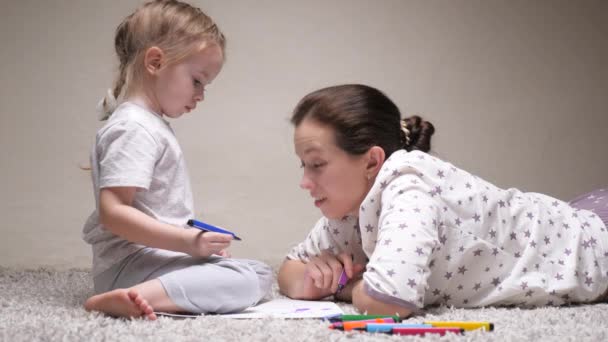 Anne, dadı, çocuk kıza resim yapmayı öğretiyor. Mutlu aile evde yerde birlikte oynuyor. Anne kızının kağıt üzerine resim çizmeyi öğrenmesine çok renkli kalemler ve keçeli kalemlerle yardım ediyor.. — Stok video