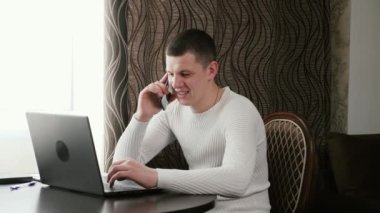Genç bir iş adamı, iş adamı, dizüstü bilgisayarla çalışıyor, akıllı bir telefonla konuşuyor. Ofiste, evde bilgisayar ve cep telefonu kullanan profesyonel bir erkek. Modern dizüstü bilgisayarla çalışan serbest çalışan biri..