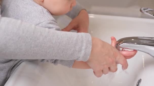 Un bambino piccolo si lava le mani con sua madre nel bagno sopra il lavandino. Una madre aiuta suo figlio a lavarsi le mani a casa. Il ragazzo impara a lavarsi le mani da solo. Giochi l'acqua con il bambino. — Video Stock