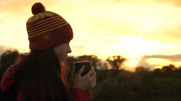 En kvinnlig resenär håller en mugg varmt kaffe i sina händer och tittar på solnedgången och vilar. Gratis flicka resenär beundrar landskapet. En turist dricker te från en mugg i solen. Resor, vandring — Stockvideo