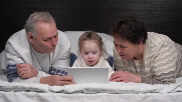 Lycklig familj, moderna morföräldrar leker med ett barn, en flicka som ligger med en surfplatta online hemma. Barn, flicka visar något i tabletten för morföräldrar. Äldre par med barnbarn och pryl — Stockvideo
