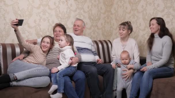 Ευτυχισμένη μεγάλη οικογένεια διαφορετικών γενεών φωτογραφίζεται στο smartphone, βγάζοντας selfie μαζί στο σπίτι. Ένα ηλικιωμένο, παντρεμένο ζευγάρι τραβάει βίντεο στο τηλέφωνο, παιδιά, εγγόνια, δισέγγονα. — Αρχείο Βίντεο