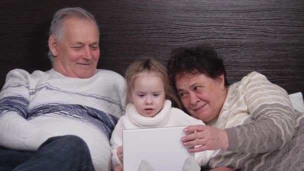 Familia feliz, abuelos modernos juegan con un niño, una niña acostada con una tableta en línea en casa. Niño, niña muestra algo en la tableta a los abuelos. Pareja mayor con nieta y gadget — Vídeos de Stock