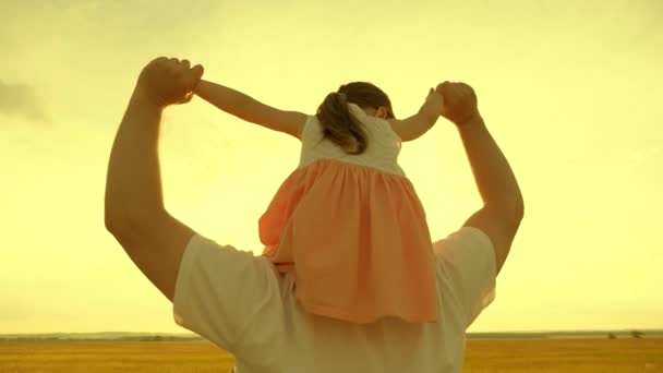 Ojciec chodzi z córką na ramionach w promieniach zachodzącego słońca. Szczęśliwa rodzina odpoczywa w parku. Tata nosi swoje ukochane dziecko na szyi na słońcu. Dziecko z rodzicem spaceruje w weekend. — Wideo stockowe