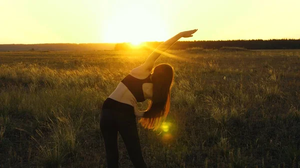 Free young woman goes in for sports in summer park at sunset. A healthy beautiful girl is engaged in fitness, outside city in sun. Workout and warm-up in outdoors. Girl breathes fresh air on field.
