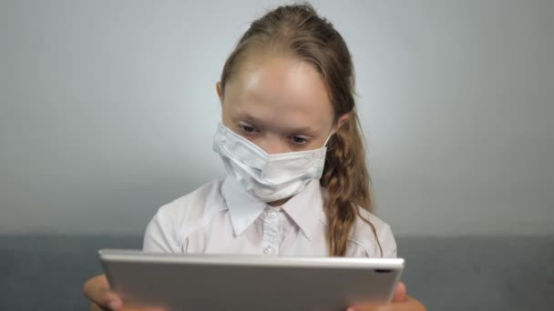Uno studente con una maschera medica insegna i compiti usando un tablet. Una ragazza con una maschera protettiva durante una lezione online. Un bambino in maschera gioca sul suo tablet nella stanza sul divano. Quarantena, pericolo virale — Video Stock
