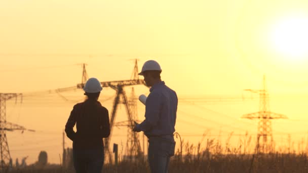 Bir grup enerji mühendisi açık havada bir çalışma planı üzerinde çalışıyor. Mühendis erkek ve kadın gün batımında elektrik kuleleriyle sahada çalışıyor. Mühendislerin takım çalışması. Yüksek voltajlı elektrik santrali işletme projesi. — Stok video