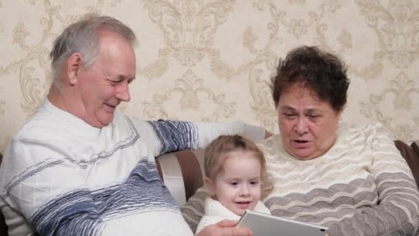 Abuelo, la abuela está jugando con el niño juntos en el gadget moderno en la habitación. Niño, niña muestra algo en la tableta a los abuelos. Una pareja de ancianos con nieta están utilizando tableta en línea — Vídeos de Stock