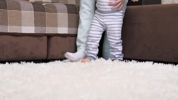Happy mom holding hand helps cute little baby take first steps at home. The concept of childhood and a happy family. Infant barefoot girl boy learns to walk while standing on a warm floor. — Stock Video