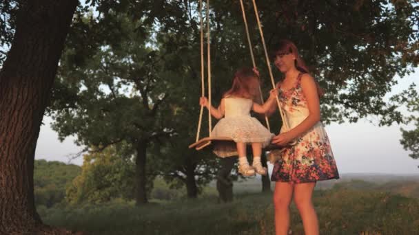 Happy family, young mom and little daughter on a swing in sunlight in park. A child, a girl plays with her mother, on wooden swing on playground. Baby and mother relaxing outdoors, happy childhood — Stock Video