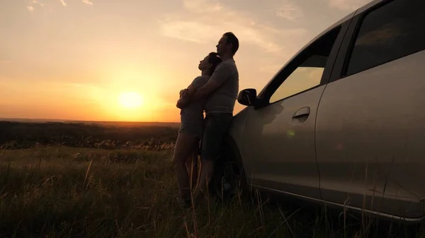 Tourists travel by car, hug, admire sunrise, nature. Free travelers, tourists. Happy enamored travelers man and woman stand next to car and admire beautiful sunset at campsite. Family travel by car.