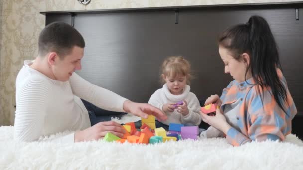 Los padres, papá, mamá, hija juegan con cubos en la guardería en la cama. Enseñando a los niños a través del juego. Niño, padre y madre están jugando a construir una casa familiar. Familia feliz. Juegos educativos para niños. — Vídeo de stock