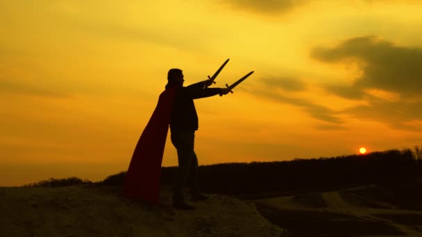 A winner with swords in his hand and in a red cloak stands on a mountain in the sunset light. free man plays a superhero. game of the Roman legion. free male knight prays with outstretched swords. — Stock Video