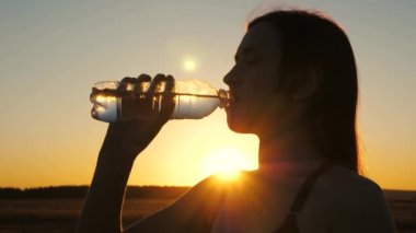 Genç bir kadın idmandan sonra şişeden su içer. Gün batımında dışarıda koşuyor. Yazın, ilkbaharda parkta açık hava sporları. Kız antrenmandan sonra temiz, serin maden suyu içiyor..