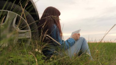 Kadın kamp alanında durmuş, tabletle dinleniyor, doğa. Kız arabanın yanında dinleniyor, rotayı kontrol ediyor. Tablet bilgisayarlı gezgin sürücü kamp alanında durdu. Araba, açık hava eğlencesi.