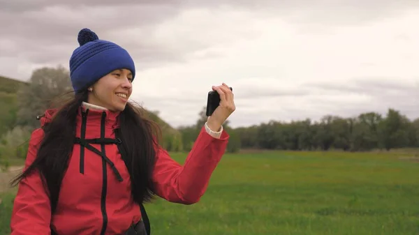 Gratis Giovane ragazza blogger turistica registra video selfie ai piedi delle montagne utilizzando uno smartphone con un bellissimo paesaggio in background. Donna sana allegra viaggia e fotografa la natura per telefono. — Foto Stock