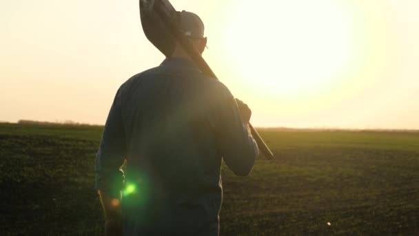A farmer with a shovel in his hands walks across the field with a shovel in the sun. An agronomist walks on a black fertile plowed land at sunset. Worker with a shovel in a field at sunset. — Stock Video