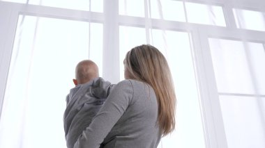 Kid son and young mom are playing looking out the window together waiting for dad. Happy family. Mother, child, daughter, play and look out into the street, accompanying the father. Infant and mommy.
