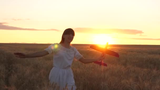 太陽に向かって小麦畑を走っている子供の女の子の手の中に飛行機。飛行についての航空会社の女の子、夢や空想。幸せな子供時代。飛行機に乗って世界を旅する10代の夢. — ストック動画