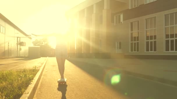 Heureuse fille à la mode voyage dans les rues de la ville en rayons de soleil sur une planche à roulettes. Gratuit adolescent fille apprend à monter skateboard sur rue au soleil. Happy skateboarder promenades sur la route en ville au coucher du soleil. — Video