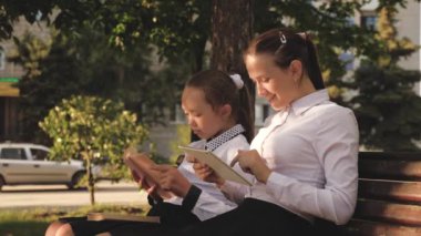 Girls School Girls are sitting on bench in park, holding textbooks and tablet. Teenagers in schoolyard in spring. Girls teach outdoor lessons, modern, traditional education. Smart, educated, children
