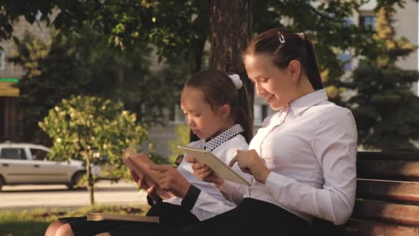 Ragazze Scuola Ragazze sono seduti sulla panchina nel parco, in possesso di libri di testo e tablet. Adolescenti nel cortile della scuola in primavera. Le ragazze insegnano lezioni all'aperto, istruzione moderna e tradizionale. Intelligenti, istruiti, bambini — Video Stock