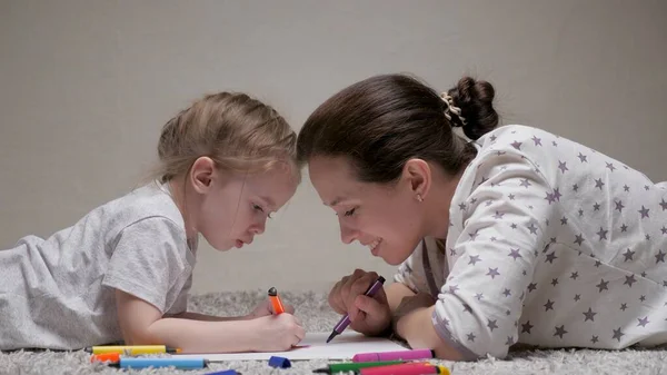 Child and Mom, nanny, teaches girl to draw. Happy family playing together at home on floor. A mother helps her daughter learn to draw on paper, coloring with multi-colored pencils and felt-tip pens. — Stock Photo, Image