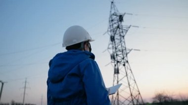 Beyaz kasklı bir kadın güç mühendisi tabletteki elektrik sensörlerinden elde edilen verileri kullanarak güç hattını kontrol ediyor. Gün batımında yüksek voltajlı elektrik hatları. Dağıtım ve elektrik tedariği. temiz enerji
