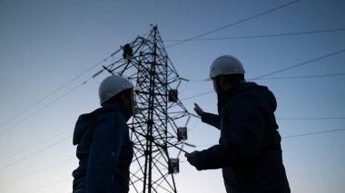 Elektrik mühendisleri çalışma planını tartışıyorlar. Gün batımında elektrik kuleleri olan iki mühendis sahada duruyor. Mühendislerin silueti yüksek voltajlı elektrik üretimine bakıyor. Mühendislik ekibi.
