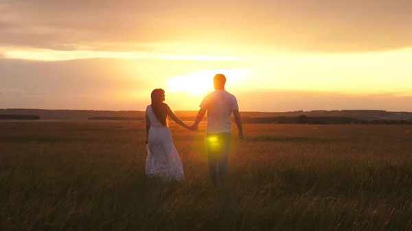 Une belle fille et un mec marchent main dans la main dans le champ, au soleil sur l'herbe verte. Silhouette d'homme et d'une femme au coucher du soleil. Un jeune couple amoureux voyage. Date romantique et amour dans la nature. — Photo