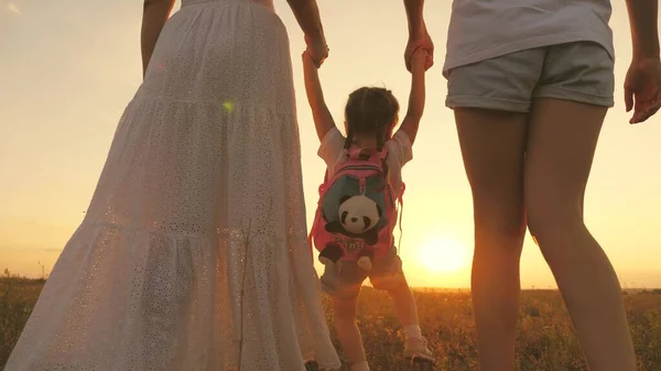 Šťastná rodina, matka, dítě a starší dcera se procházejí v parku při západu slunce. Veselé dítě se drží za ruce matky a sestry a poskakuje v paprscích slunce. Děti a rodiče chodí na jaře — Stock fotografie