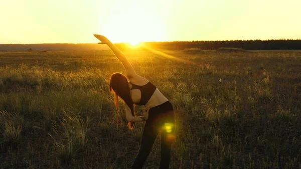 Gratis jonge vrouw gaat sporten in het zomerpark bij zonsondergang. Een gezond mooi meisje is bezig met fitness, buiten de stad in de zon. Trainen en opwarmen in de buitenlucht. Meisje ademt frisse lucht op het veld. — Stockfoto