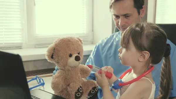 Opiekuńczy profesjonalny pediatra bawiący się w biurze z małym dzieckiem. Dziewczynka bawi się pluszową zabawką słucha go stetoskopem. dziecko pacjent siedzi na kolanach pediatry. — Zdjęcie stockowe