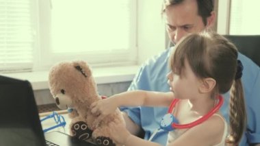 Ofiste küçük bir çocukla oynayan profesyonel bir erkek çocuk doktoru. Küçük kız oyuncak ayıyla oynuyor ve steteskopla onu dinliyor. Yeni yürümeye başlayan hasta çocuk doktorunun kucağına oturuyor..