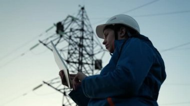 Beyaz kasklı kadın güç mühendisi güç hattını kontrol ediyor tabletteki bilgisayarla, güç sisteminin uzaktan kumandasıyla. Gün batımında yüksek voltajlı elektrik hatları. Dağıtım ve elektrik tedariği. Enerji