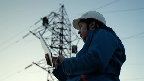 Vrouwelijke power engineer in witte helm controleert de stroomleiding met behulp van de computer op tablet, afstandsbediening van het energiesysteem. Hoogspanningsleidingen bij zonsondergang. Distributie en levering van elektriciteit. Energie — Stockvideo