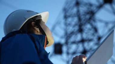 Elektrik mühendisi beyaz kasklı bir kadın güç hattını bilgisayar tableti kullanarak kontrol ediyor. Dışarıda çalışan kadın işçi. Gün batımında yüksek voltajlı elektrik hatları. Elektrik dağıtımı ve tedariği.