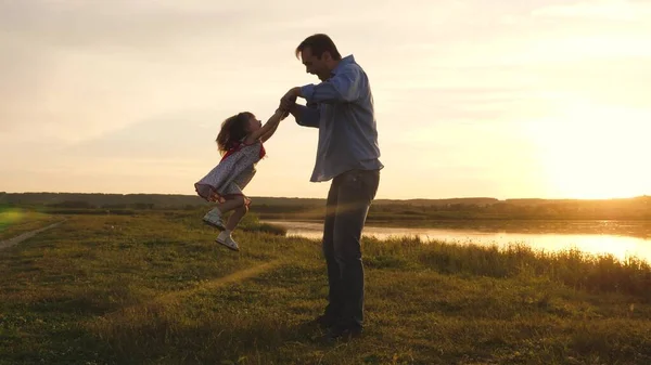 Mała córeczka bawi się z tatą w parku na słońcu. Tata uściska dłonie szczęśliwemu dziecku w promieniach zachodu słońca w parku. Szczęśliwa rodzina na wakacjach. Wesoły dzieciak bawi się z ojcem.. — Zdjęcie stockowe