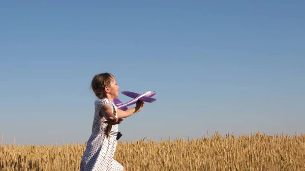 幸せな女の子は青い空の下の小麦畑でおもちゃの飛行機で走っています。子供はおもちゃの飛行機で遊ぶ。10代の頃はパイロットになる夢を見ました。少女はパイロットとなり宇宙飛行士になりたいのです — ストック写真