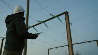 Koruyucu beyaz miğferdeki güç mühendisi güç hattını kontrol etmek için bilgisayar tableti ve güç sistemine uzaktan erişim kullanarak kontrol ediyor. Gün batımında yüksek voltajlı elektrik hatları.